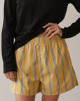 A person in a black long-sleeve shirt holds the waistband of The Pop Boxer by Donni, featuring relaxed-fit yellow shorts with blue and white stripes. Made from breathable cotton poplin, these shorts contrast with the plain, neutral background.