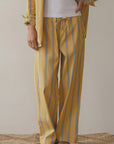 The person, showcasing a versatile style for relaxed comfort, is dressed in loose-fitting mustard yellow "The Stripe Pop Drawstring Pant" by Donni, made of cotton poplin. They pair it with a matching top over a white shirt, stand on a woven rug, and wear brown slippers.