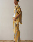 A relaxed individual with tied-up brown hair wears a yellow striped shirt and Donni's The Stripe Pop Drawstring Pant made of cotton poplin over a white top. They stand barefoot on a textured surface against a plain, light background.