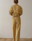 A person with hair in a bun, seen from behind, wears a yellow and white striped outfit by Donni, featuring soft cotton poplin with long sleeves and The Stripe Pop Drawstring Pant. The pants have a comfy drawstring waist that enhances the woven texture against the plain backdrop.