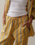 Donni's The Stripe Pop Drawstring Pant, in soft cotton poplin with blue and white stripes, offers comfort with its drawstring waist. Paired with a partially unbuttoned white ribbed tank top, this relaxed yellow pajama set allows easy hand-tucking into pockets.