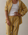 A person wears Donni's The Stripe Pop Drawstring Pant, featuring a loose yellow design with blue and white vertical stripes. Paired with a white tank top, their left hand is on their hip while the right is relaxed by their side, exuding casual elegance in cotton poplin.