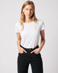 A person with long blonde hair wears the Majestic Filatures Deluxe Cotton S/S Crewneck and high-waisted black pants, standing against a light grey background with hands in pockets and looking at the camera.