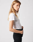 A woman with long blonde hair wears a white Majestic Filatures Deluxe Cotton S/S Crewneck tee and black pants, standing in profile against a plain light background, looking seriously at the camera.