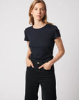 A person with long, light brown hair stands against a plain background, wearing Majestic Filatures’ Deluxe Cotton S/S Crewneck in black and black pants, hands in pockets, looking confidently at the camera.