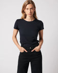 A person with long blonde hair wears the Majestic Filatures Deluxe Cotton S/S Crewneck in black, paired with black pants, standing hands-in-pockets and facing the camera against a plain light background.
