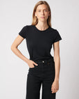 A person with long light brown hair wears the Majestic Filatures Deluxe Cotton S/S Crewneck and black jeans, standing against a plain light background, looking directly at the camera with a neutral expression and one hand in their pocket.