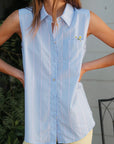 A person wearing a Donni light blue striped sleeveless cotton poplin shirt and pale yellow pants, with hands on hips, only the torso and legs visible.