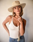 A woman with short blonde hair wearing a wide-brimmed Ninakuru Paloma Straw Hat and a white tank top, pensively touching her chin, against a neutral backdrop.