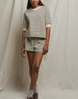 A woman stands before a light fabric backdrop, wearing the Perfectwhitetee French Terry Short set with a short-sleeve top, gray socks, and brown shoes.