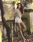 A woman with long curly hair leans on a stone balustrade in a garden, wearing the DOEN Iona Short and a light knit cardigan, surrounded by fallen leaves and trees.