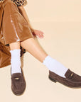 A person sits cross-legged on a cream background in a brown leopard-print top, shiny brown skirt, white ribbed socks, and Freda Salvador's Elba Espresso Suede Loafer.