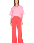 A person with straight brown hair and a neutral expression stands against a white background, wearing a loose pink T-shirt, Xirena Emmette Sweatpant in bright red, and pink shoes.