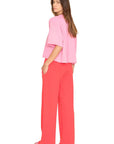 A woman with long brown hair and a neutral expression stands against a white background, wearing the Xirena Emmette Sweatpant—an ultra-soft fleece wide leg style—paired with a loose-fitting pink top.