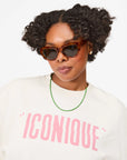 A woman with natural curly hair wears Clare Vivier’s Sophia Sunglasses—brown cat-eye frames made from recycled material—a green beaded necklace, and a white shirt with bold pink “ICONIQUE,” facing the camera on a plain white background.