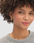 A woman with short curly hair wears a light grey sweater and Clare Vivier Etoile Hoops, smiling softly against a neutral background.