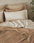 A neatly made bed features patterned beige and white pillows, a striped pillow, and beige and pink bedding. The Coyuchi Inc Mirada Matelasse Blanket in organic cotton adds comfort. A potted green plant sits by the bed against a light wall.