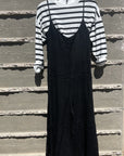 A black Linen Jumper over a white and black striped long-sleeve shirt, hanging on a hanger against a textured gray concrete wall in Scottsdale, Arizona by Importations Eternelle Inc.