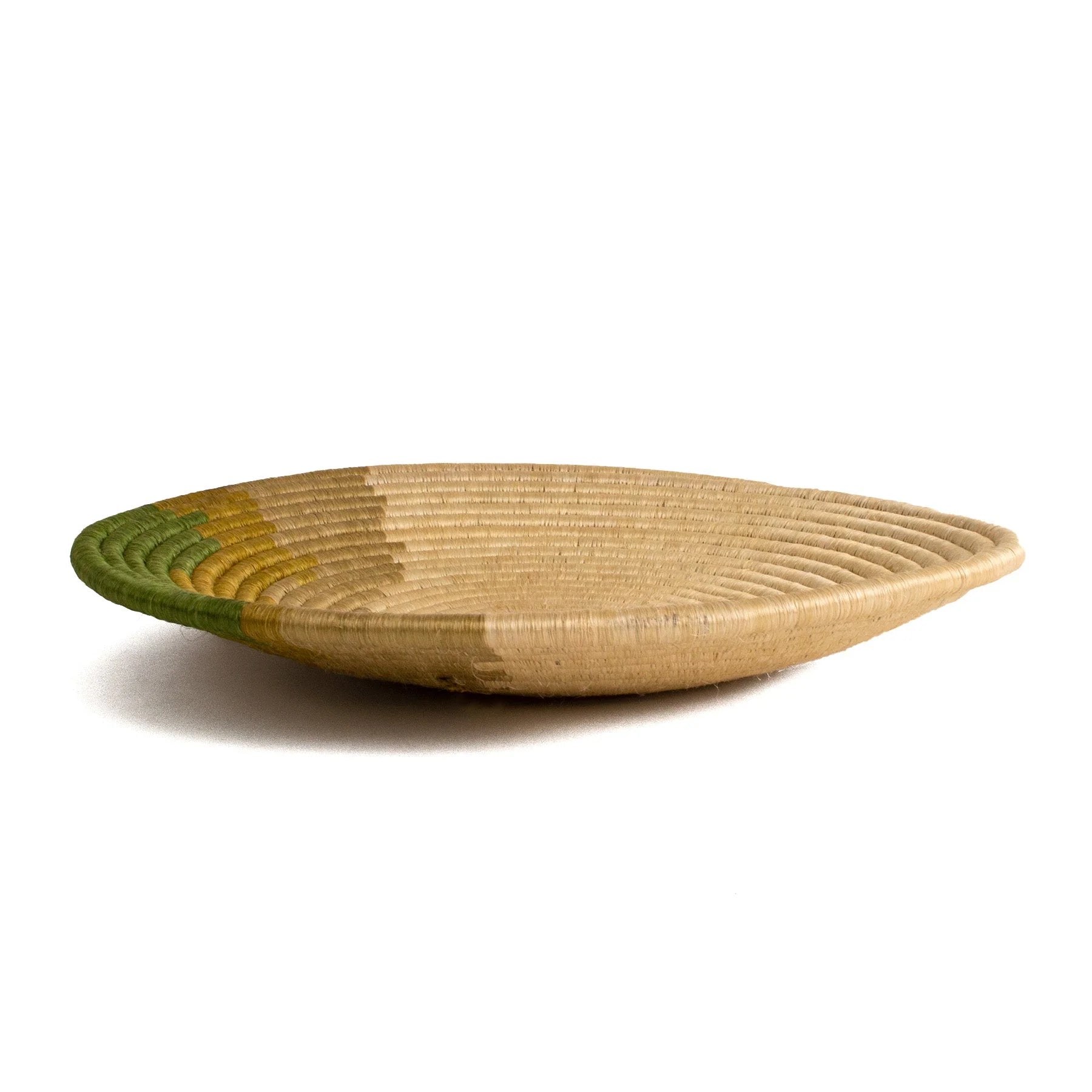 A Restorative Woven Bowl 21" Moss by Kazi Goods, a shallow, oval-shaped woven basket in natural tan color with subtle green accents radiating outward from the center is showcased against a plain white background. This piece reflects the tranquil aesthetic of a Scottsdale Arizona bungalow.
