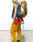 A person stands sideways against a plain white background, wearing the Free City Out of Sight Poplin Jump Pant in yellow, a black cap, denim vest, green sneakers, and carrying a large yellow and gray backpack.