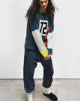 A person with long dark hair wears the Free City (sparrow, LLC) CanyonKid 72' Cashmere Lux Crew sweater, loose blue sweatpants, white socks, and black slippers while standing indoors on a concrete floor against a plain background.