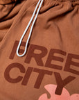 Close-up of FREECITY Large Sweatpant by Free City (sparrow, LLC) in brown French terry with white drawstrings. Features bold “FREE CITY” print and partial pink graphic near the hem. Made in USA for comfort and style.