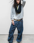 A woman with long dark hair poses against a plain background, wearing Free City Loose Drawcord Biggy Pant, a loose grey sweatshirt, black scarf, and green sneakers. She touches her head as she looks at the camera.