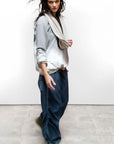 A person with long dark hair wears Free City Loose Drawcord Biggy Pant, a light gray sweatshirt, green sneakers, and a beige scarf while standing on a concrete floor against a plain white wall, looking at the camera.
