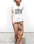 A person with short blonde hair wears a white "FREE CITY" t-shirt, Free City Loose Drawcord Biggy Pant, green sneakers, and layered necklaces, standing on a gray concrete floor against a plain white wall.
