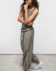 A woman with long wavy hair stands against a white background wearing a handmade FreeCity SuperVintage Tank by Free City (sparrow, LLC), loose gray drawstring pants, white sneakers, and layered necklaces.