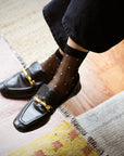 A person wearing Unified Sheer Dot Socks—stylish sheer black women's socks—and black loafers with gold hardware, sits cross-legged on a light-colored carpeted floor.