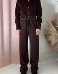 A person stands indoors in a dark brown button-up shirt and Frank & Eileen’s Catherine Long Sweatpant, made from 100% cotton with a waist tie. They wear black shoes, with pink curtains and beige pampas grass as background decor.
