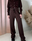 A person stands indoors wearing Frank & Eileen’s Catherine Long Sweatpant, paired with a dark brown corduroy button-up shirt and black shoes. Light pink curtains and dried grasses complete the background.