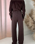 A person stands facing away, wearing Frank & Eileen’s Catherine Long Sweatpant with a dark brown long-sleeve shirt. Beige pampas grass and pink curtains create a soft backdrop.