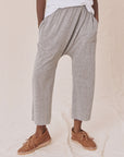 Against a cream fabric backdrop, someone wears The Great Inc.'s gray Jersey Crop harem pants with a dropped rise and brown moccasin-style shoes. The loose-fit design, similar to wider straight leg pants, pairs hands in pockets with a relaxed white top.
