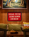 A cozy room with wood-paneled walls features an Oxford Pennant "Sugar We're Going Down Swingin'" 16x24 red flag above a sofa with green pillows and a zigzag cushion. A landscape painting hangs above the flag.