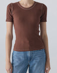 A person wearing the Cherish Tee by AMO Denim—a fitted, ribbed cotton tee with short sleeves and white seam stitching—paired with blue jeans, stands against a plain light gray background.
