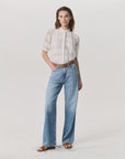 A woman stands against a plain background wearing Rag & Bone’s Logan Pant in light indigo featherweight denim, paired with a white semi-sheer blouse, brown belt, and brown sandals. One hand is in her pocket.