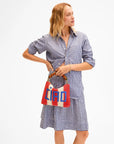 A woman in a blue and white striped button-down and gingham skirt holds the Clare Vivier Flat Clutch W/ Tabs S25, a red, cream, and blue crocheted cotton bag with bamboo handles, while looking to the side against a plain white background.