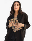 A woman with long dark hair wears a black blazer and holds the Clare Vivier Flat Clutch with Tabs 26, featuring a tan-and-black snakeskin pattern, as she poses against a plain light background.