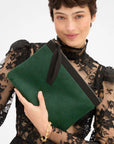 Green Clare Vivier Flat Clutch with tabs, black zipper, and wrist strap held by person in black lace top.