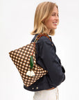 A smiling blonde woman wears a dark jacket and carries the Clare Vivier Flat Clutch with Tabs 26, featuring a handwoven leather design and a green and white flower charm on her shoulder.