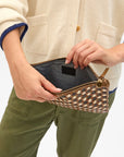 A person in a cream sweater and green pants holds the Clare Vivier Flat Clutch with Tabs 26, an open, handwoven leather zippered pouch, revealing its dark fabric interior.