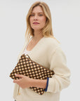 A woman with blonde hair, dressed in a cream cardigan, holds the Clare Vivier Flat Clutch with Tabs 26—a handwoven brown and black checked leather bag—against a plain, light background.