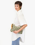 A woman with short brown hair holds a Clare Vivier Flat Clutch W/Tabs, a green and white crocheted cotton pouch with a yellow zipper, while wearing a loose white shirt over blue-and-white striped pants against a white background.