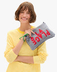 A woman with short brown hair in a yellow and white striped shirt smiles, holding the Clare Vivier La Plage Flat Clutch w/tabs, which features blue and white stripes and "PLACE" in red letters upside down.