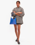 A person with arms crossed wears a black and white checkered oversized shirt, black shorts, dark sandals, and holds the Clare Vivier Foldover Clutch W/ Tabs 26 in bright blue Italian nappa leather against a plain white background.