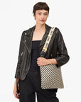 Clad in a black leather jacket and pants, someone models a patterned shoulder bag from Clare Vivier featuring the Crossbody Strap S25 with beige cotton webbing. The strap, repeatedly emblazoned with "CIAO," rests comfortably across their shoulder against a plain white backdrop.