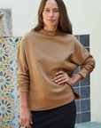 A woman with long brown hair poses indoors in the EFFIE L/S Capelet by Frank & Eileen, paired with a black skirt. She stands in front of a vibrant tiled wall, one hand on her hip.