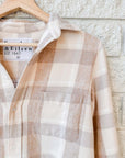 A cream and light brown plaid Silvio Untuckable Shirt by Frank & Eileen, featuring a chest pocket and a branded label, hangs on a wooden hanger against a white brick wall.